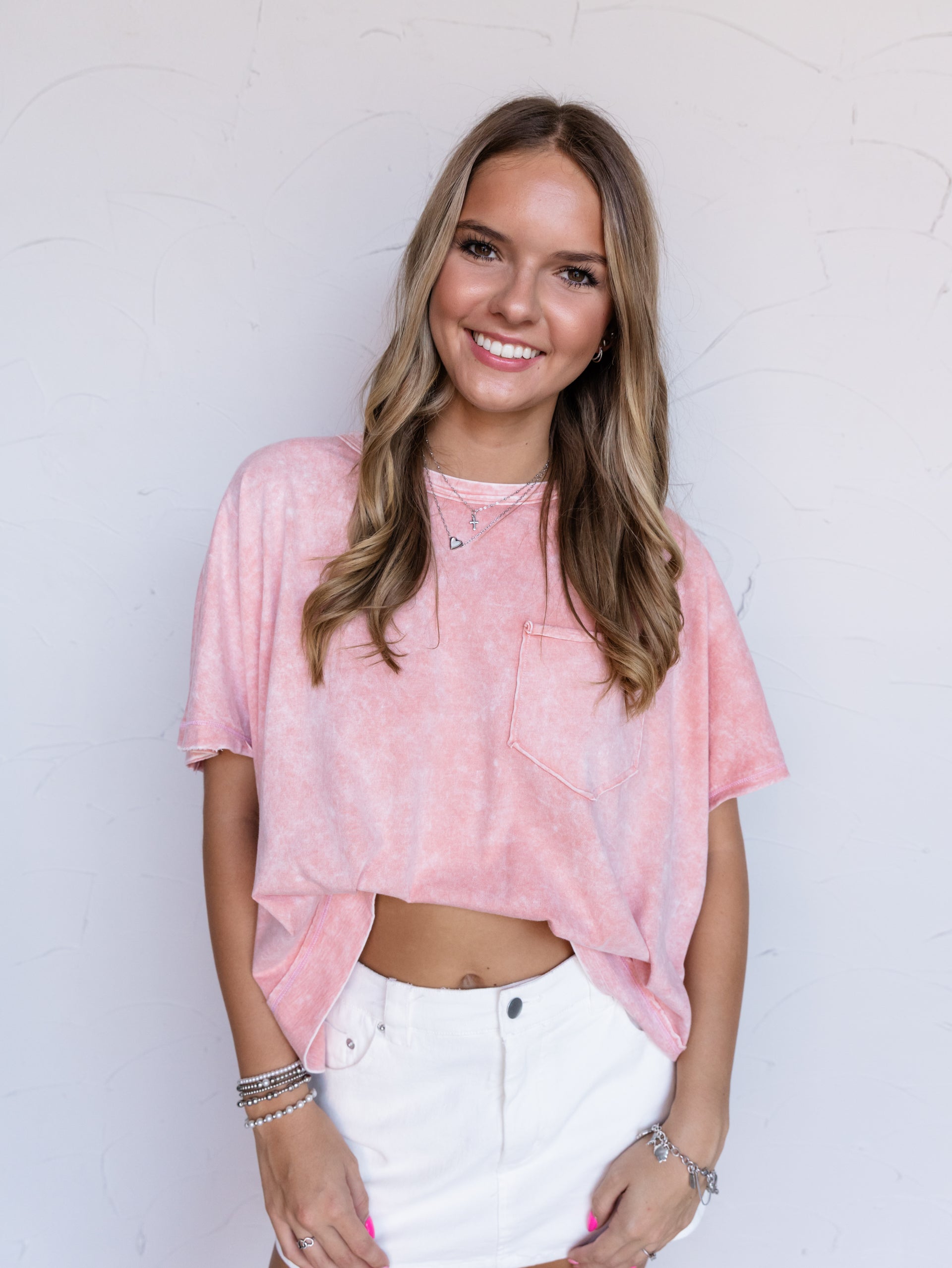 A portrait photograph focusing from the hips up of a smiling woman with long wavy blonde hair posing with her hands resting in front against a textured white wall. She is wearing an oversized pink mineral wash short-sleeved tee made from 100% cotton fabric, featuring a crew neckline, dropped shoulders, and a single chest patch pocket, styled with a high-waisted white denim miniskirt and layered silver necklaces and bracelets.