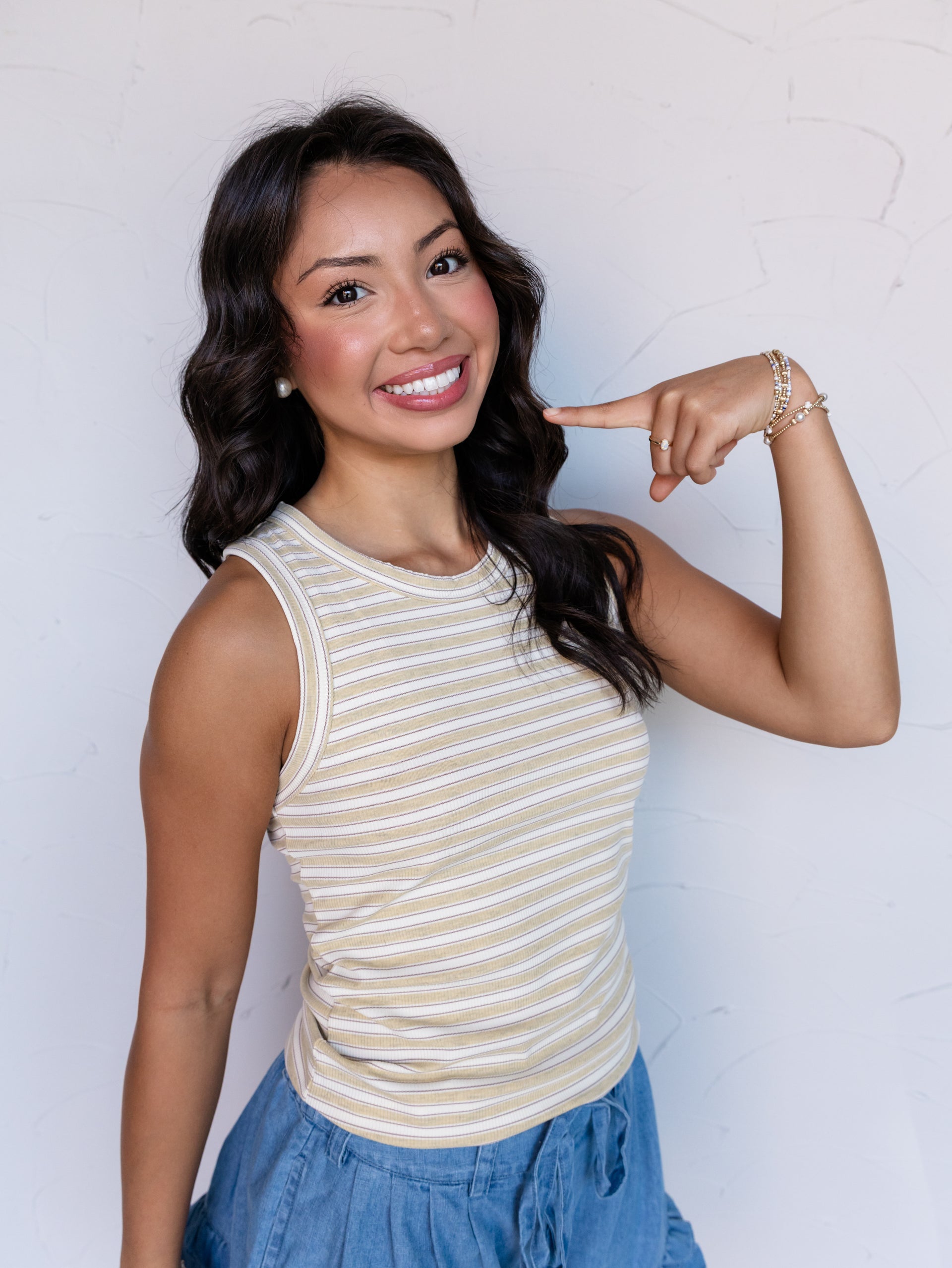 A portrait photograph focusing from the waist up of a smiling woman with shoulder-length wavy dark hair pointing to her cheek against a textured white wall. She is wearing a sleeveless ribbed tank top with thin sage green and cream horizontal stripes made from a 95% polyester and 5% spandex blend fabric, featuring a crew neck and rounded hemline, styled with chambray ruffled drawstring shorts, beaded stack bracelets, and a small ring.