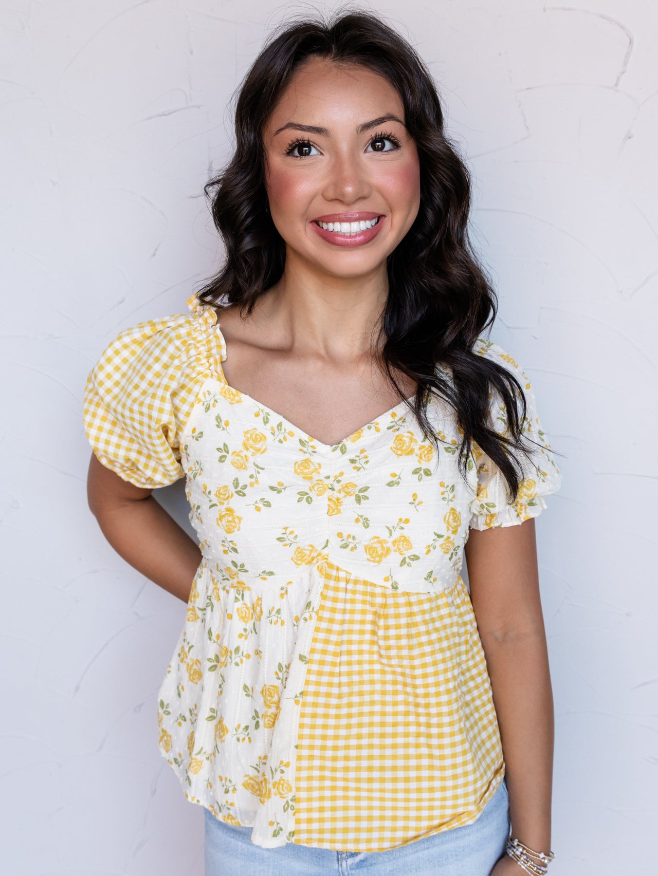 A portrait photograph focusing from the hips up of a smiling woman with shoulder-length wavy dark hair posing against a textured white wall. She is wearing a mixed-pattern short-sleeved babydoll top made from 100% cotton fabric, featuring alternating panels of yellow-and-white gingham check and a yellow floral print on white eyelet fabric, styled with a sweetheart neckline, puff sleeves, a denim miniskirt, and stacked bracelets.