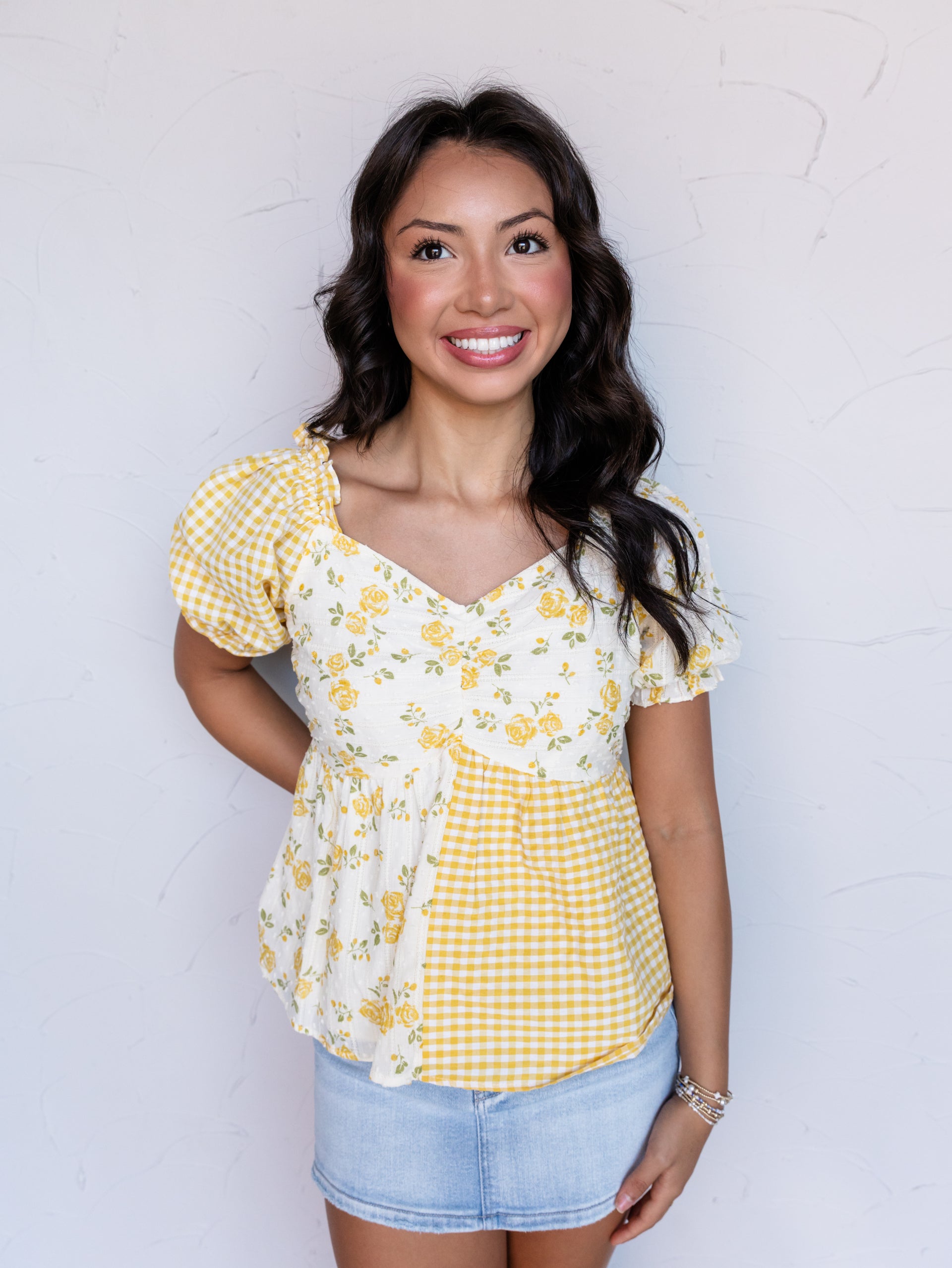 A portrait photograph focusing from the hips up of a smiling woman with shoulder-length wavy dark hair posing against a textured white wall. She is wearing a mixed-pattern short-sleeved babydoll top made from 100% cotton fabric, featuring alternating panels of yellow-and-white gingham check and a yellow floral print on white eyelet fabric, styled with a sweetheart neckline, puff sleeves, a denim miniskirt, and stacked bracelets.