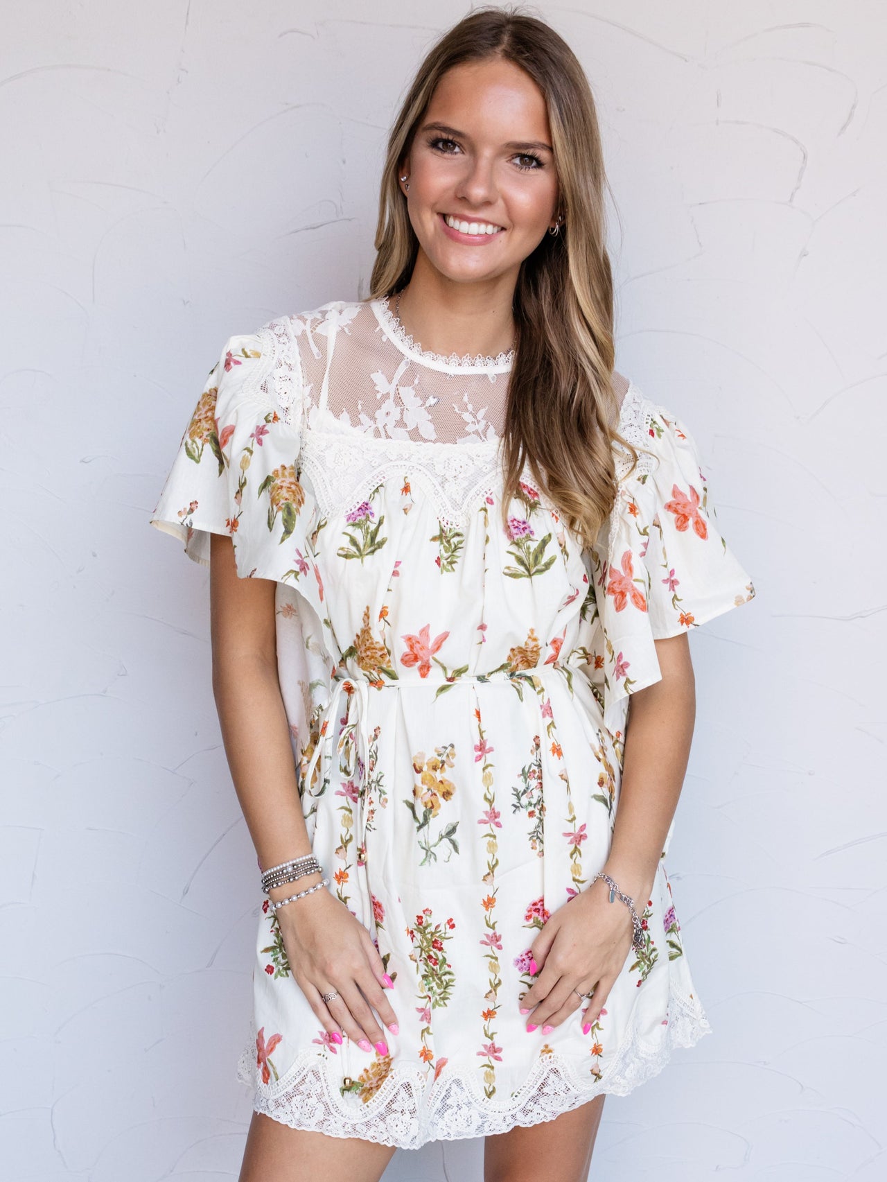 A woman modeling a short cream lace dress with a colorful floral print, standing against a white stucco wall.