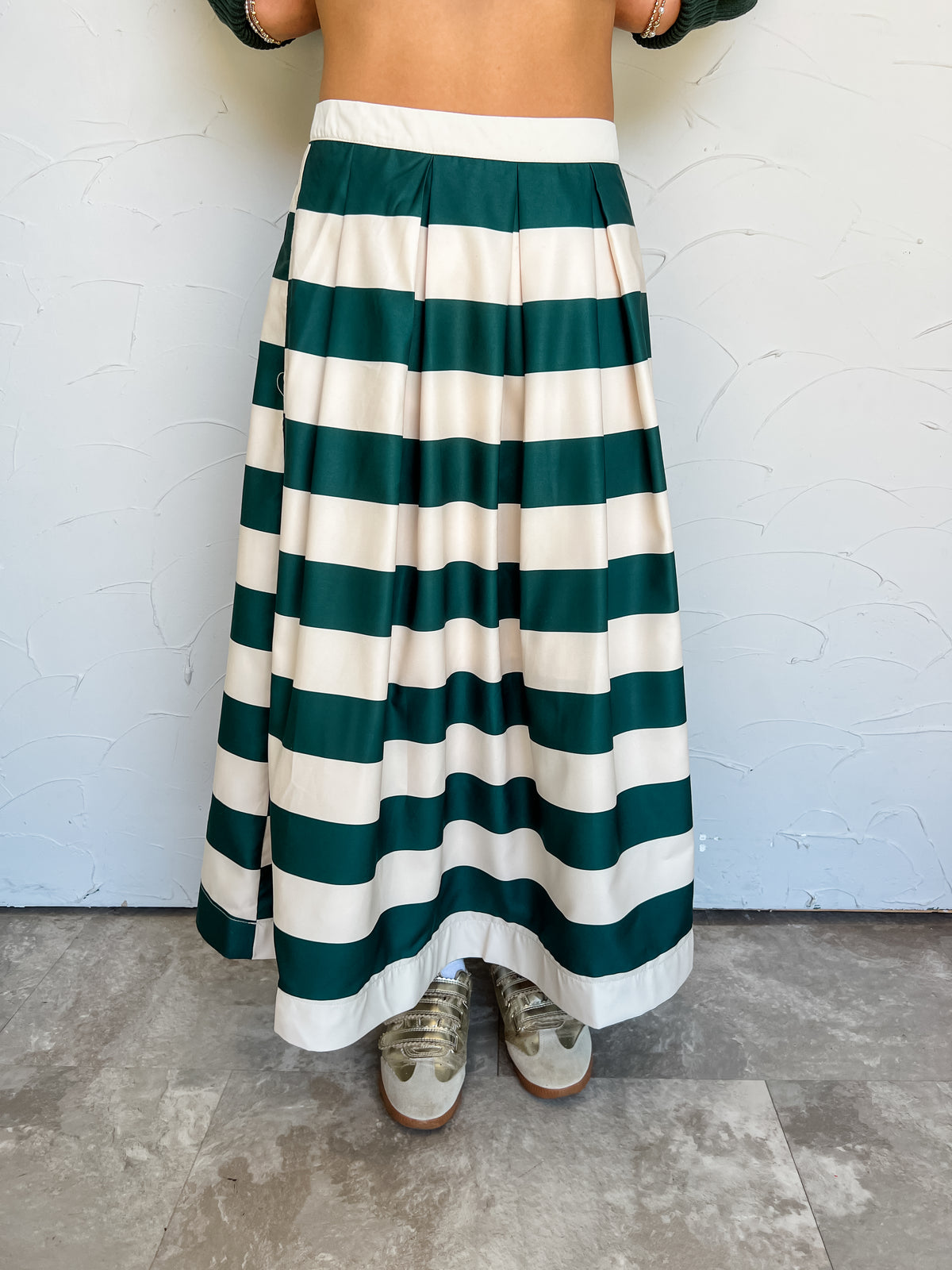 Emilia modeling the Glam My Gift To You Pleated Striped Midi Length Skirt, made of 100% polyester, featuring wide green and cream horizontal stripes, a solid cream waistband, and flowing pleats, styled with metallic sneakers.