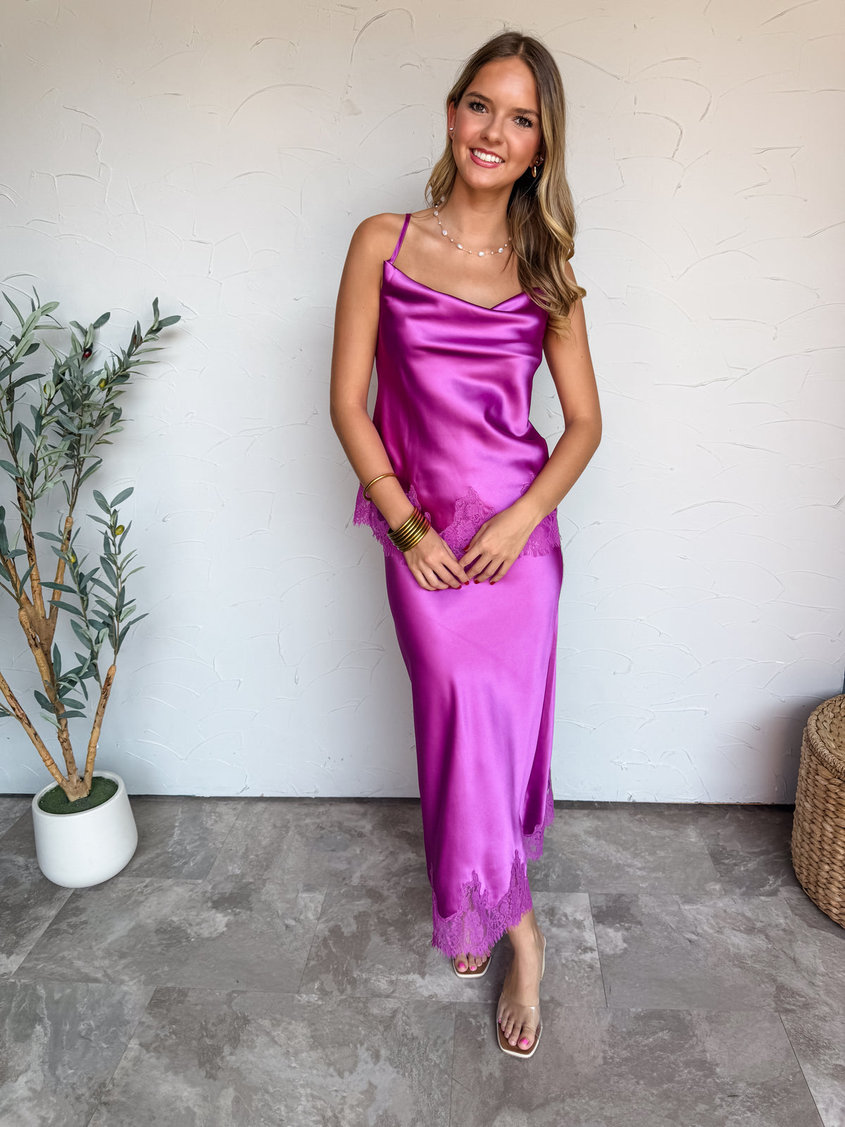 A full-length photograph of a smiling blonde woman with long wavy hair standing in front of a textured white wall next to a potted olive plant. She is wearing a two-piece vibrant magenta purple satin set made from 100% polyester, including a spaghetti strap camisole top with a draped cowl neck and eyelash lace trim at the hem, paired with a matching high-waisted bias-cut maxi skirt with lace trim along the bottom edge, styled with clear slide heels, a delicate pearl necklace, small hoop earrings, and stacke
