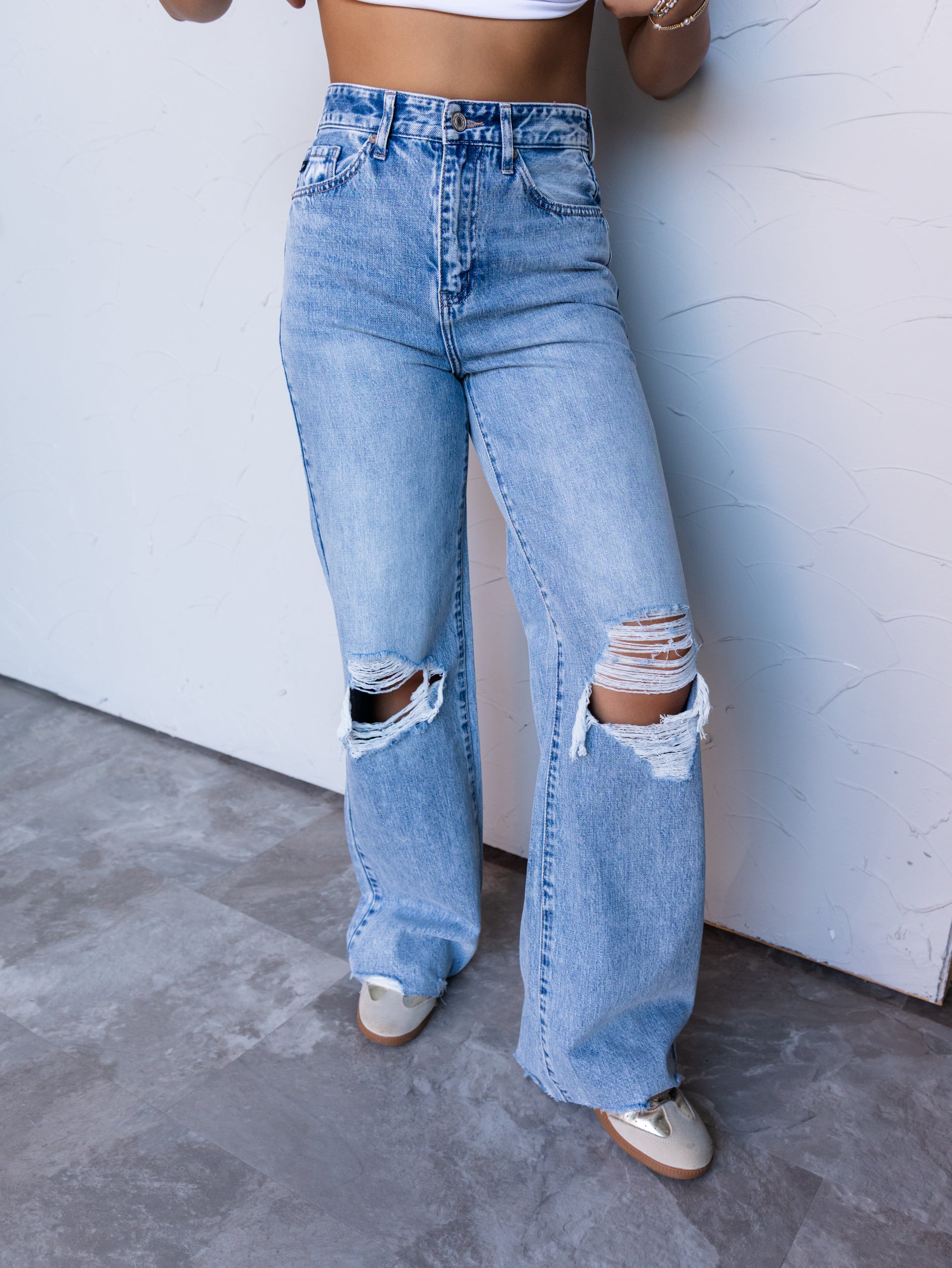 A photograph focusing from the waist down of a person posing in front of a textured white wall. They are wearing light-wash, high-waisted wide-leg denim jeans made from 100% cotton fabric, featuring shredded distressed cutouts at both knees and raw hems, styled with low-top neutral sneakers and a white crop top.