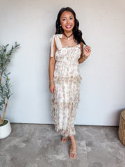 A full-length photograph of a smiling woman with long dark wavy hair holding a strand of hair while posing against a textured white wall next to a potted olive plant. She is wearing a sleeveless 100% polyester maxi dress featuring a sheer cream tulle overlay with a soft pink floral print, wide off-white ribbon ties at the shoulders, a smocked bodice, and a tiered ruffled skirt with delicate eyelash trim, styled with nude strappy heel sandals, a brown cord choker with a stone pendant, pearl earrings, and sta