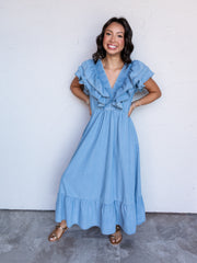 A full-length photograph of a smiling woman with shoulder-length wavy dark hair posing with her hands on her hips against a textured white wall. She is wearing a light-wash soft denim maxi dress featuring double-layered ruffle flutter sleeves, a ruched V-neck bodice, and a tiered skirt, styled with metallic gold flat sandals and stacked bracelets.