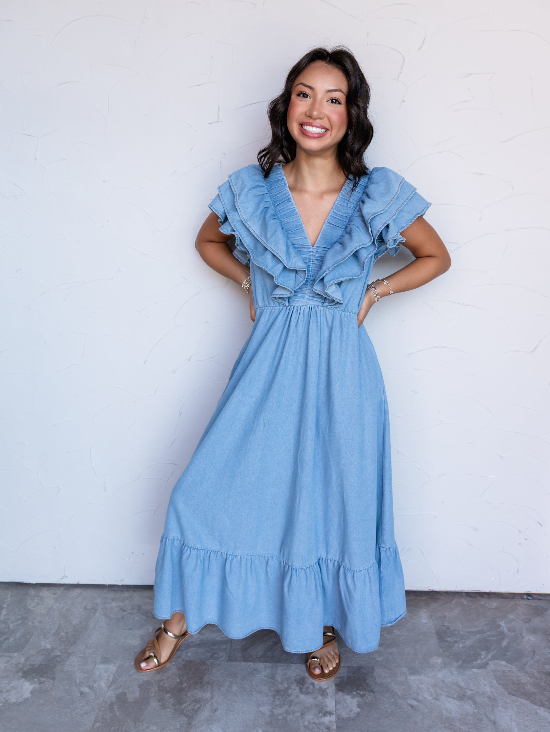 A full-length photograph of a smiling woman with shoulder-length wavy dark hair posing with her hands on her hips against a textured white wall. She is wearing a light-wash soft denim maxi dress featuring double-layered ruffle flutter sleeves, a ruched V-neck bodice, and a tiered skirt, styled with metallic gold flat sandals and stacked bracelets.