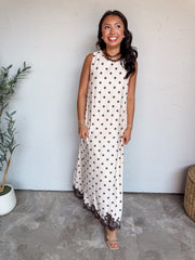 A full-length photograph of a smiling woman with long dark wavy hair looking to the side while holding the sides of her dress against a textured white wall next to a potted olive plant. She is wearing a sleeveless maxi dress made from an 81% viscose and 19% nylon blend, featuring a cream background with brown polka dots, a crew neckline, a relaxed column fit, and a dark brown scalloped lace trim along the bottom hem, styled with nude strappy sandals, a brown ribbon choker necklace with a gold pendant, bead 