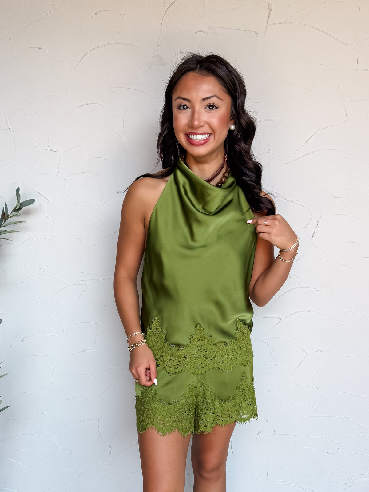 A full-length photograph of a smiling woman with dark wavy hair looking off to the side while touching her hair against a textured white wall next to a potted olive plant. She is wearing a matching two-piece olive green satin set made from 100% polyester, including a sleeveless high-neck halter top with a draped cowl neckline and scalloped lace trim along the hem, paired with matching satin shorts featuring a lace-trimmed hem, styled with nude strappy sandals, a brown cord choker, pearl stud earrings, and s