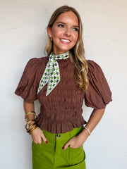 A portrait photograph of a smiling blonde woman with wavy hair looking off to the side with her hands in her pockets against a textured white wall. She is wearing a short-sleeved chocolate brown top made from a 71% rayon and 29% nylon blend, featuring full smocking through the bodice, voluminous puff sleeves, a ruffled peplum hem, and an attached green and white geometric argyle patterned neck scarf tied around the high collar, tucked into vibrant lime green trousers, styled with gold hoop earrings and stac