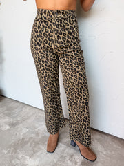 A waist-down photograph of a woman posing against a textured white wall. She is wearing high-waisted straight-leg pants made from a 97% cotton and 3% spandex blend, featuring an all-over tan, brown, and black leopard animal print, a classic button-fly closure, belt loops, and a raw-edge cropped ankle hem, styled with square-toe tan suede ankle boots with dark stacked block heels.