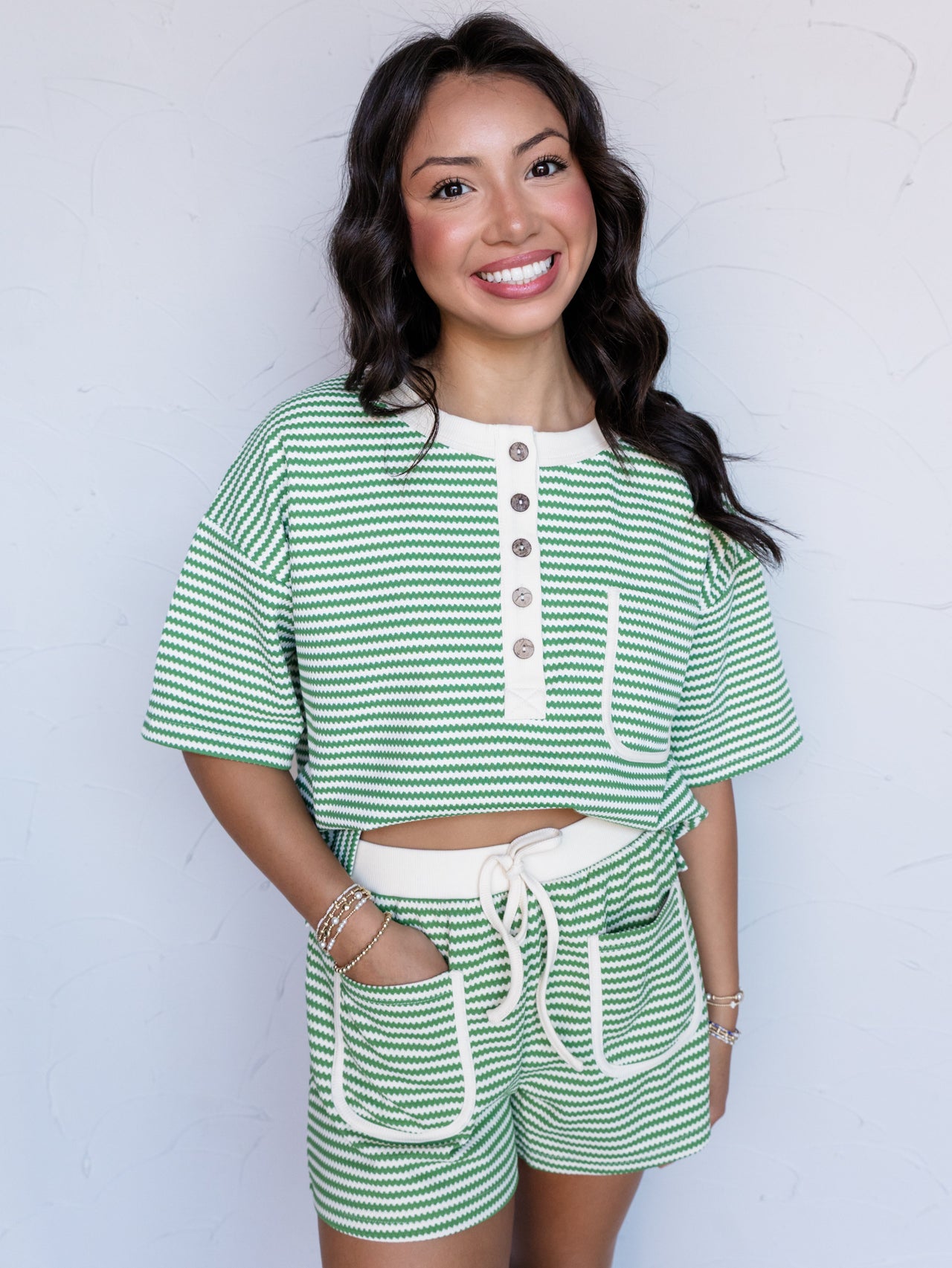 A portrait photograph focusing from the mid-thigh up of a smiling woman with dark wavy hair posing with one hand in her pocket against a textured white wall. She is wearing a matching two-piece set featuring green and cream wave-textured horizontal stripes made from a 95% polyester and 5% spandex blend fabric, consisting of a short-sleeved henley top with a cream button placket and chest pocket paired with high-waisted shorts featuring an elastic drawstring waistband and cream-trimmed front patch pockets.