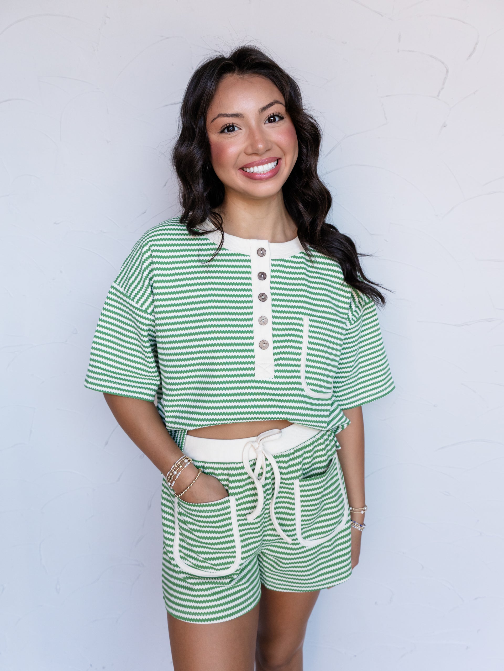 A portrait photograph focusing from the mid-thigh up of a smiling woman with dark wavy hair posing with one hand in her pocket against a textured white wall. She is wearing a matching two-piece set featuring green and cream wave-textured horizontal stripes made from a 95% polyester and 5% spandex blend fabric, consisting of a short-sleeved henley top with a cream button placket and chest pocket paired with high-waisted shorts featuring an elastic drawstring waistband and cream-trimmed front patch pockets.