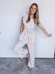 A full-length photograph of a smiling woman with long blonde hair with dark roots posing playfully with one leg bent against a textured white wall. She is wearing a taupe and white vertical pinstriped sleeveless jumpsuit made from 100% cotton fabric, featuring white eyelet lace trim along ruffle-trimmed shoulder straps, a button-down bodice, a matching tied belt at the waist, and wide-leg pants, styled with flat metallic sandals and simple jewelry.