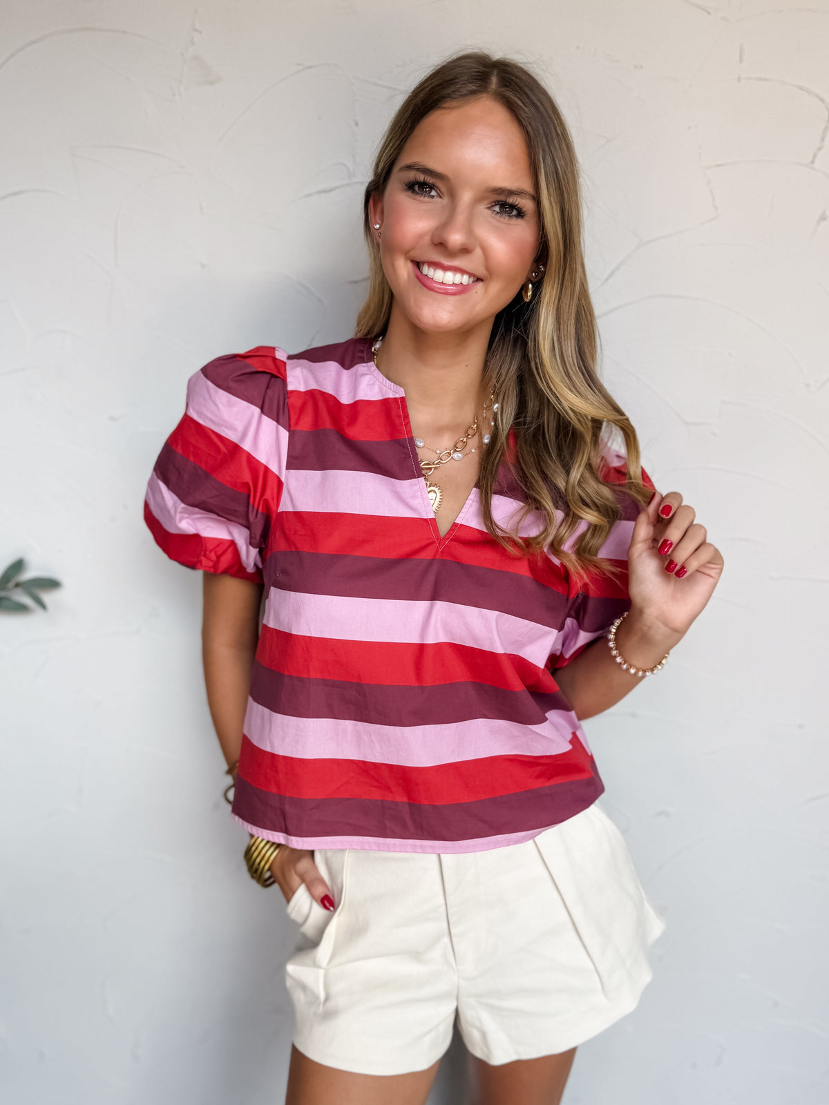 A three-quarter length photograph of a smiling blonde woman with wavy hair touching her hair while posing with one hand in her pocket against a textured white wall. She is wearing a short-sleeved 100% cotton top featuring wide horizontal stripes in red, burgundy, and light pink, designed with a split V-neckline and puffed short sleeves, paired with pleated off-white high-waisted shorts, and styled with layered gold chain necklaces, gold hoop earrings, stacked gold bangles, and a delicate beaded bracelet.
