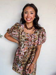 A waist-up photograph of a smiling woman with long dark wavy hair posing with one hand on her hip against a textured white wall. She is wearing a short-sleeved 100% cotton mini dress with a rich brown background covered in a detailed floral pattern featuring pink, muted yellow, and green flowers. The dress features a square neckline, small front buttons, and short puff sleeves with elastic cuffs, styled with a brown beaded choker necklace, pearl stud earrings, and stacked delicate beaded bracelets.