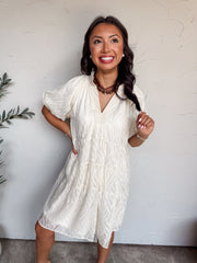 A three-quarter length photograph of a smiling woman with long dark wavy hair looking up and holding a lock of her hair while posing against a textured white wall. She is wearing a short-sleeved cream white mini dress made from 100% polyester, featuring an all-over textured wave motif, a split V-neckline with a subtle ruffle trim along the collar, puff sleeves, and a tiered swing fit, styled with a brown ribbon choker necklace with a round pendant, pearl stud earrings, and delicate beaded bracelets.