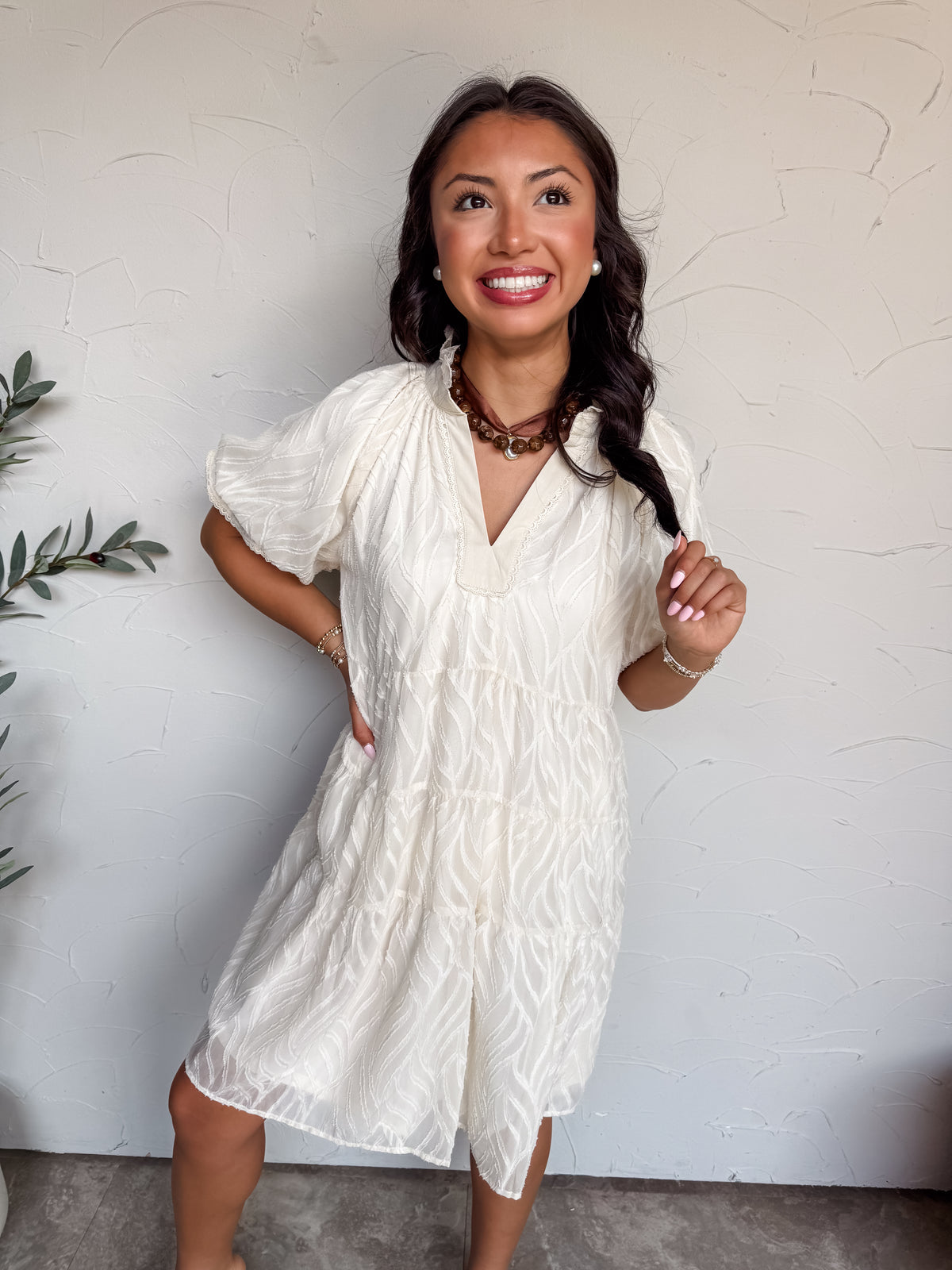 A three-quarter length photograph of a smiling woman with long dark wavy hair looking up and holding a lock of her hair while posing against a textured white wall. She is wearing a short-sleeved cream white mini dress made from 100% polyester, featuring an all-over textured wave motif, a split V-neckline with a subtle ruffle trim along the collar, puff sleeves, and a tiered swing fit, styled with a brown ribbon choker necklace with a round pendant, pearl stud earrings, and delicate beaded bracelets.