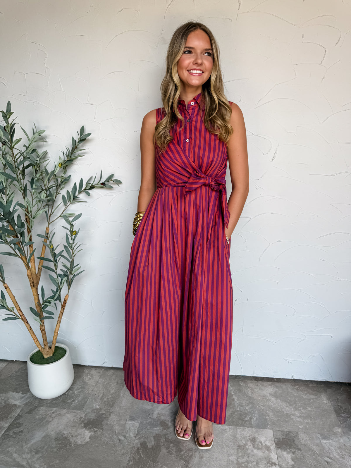 A full-length photograph of a smiling blonde woman with wavy hair posing with one hand in her pocket against a textured white wall next to a potted olive plant. She is wearing a sleeveless 100% cotton maxi dress with bold vertical red and purple stripes, featuring a pointed collar, button-up front bodice, an adjustable waist tie knot, side pockets, and an ankle-length flared skirt, styled with white flat sandals, stacked gold bangles, and delicate bracelets.