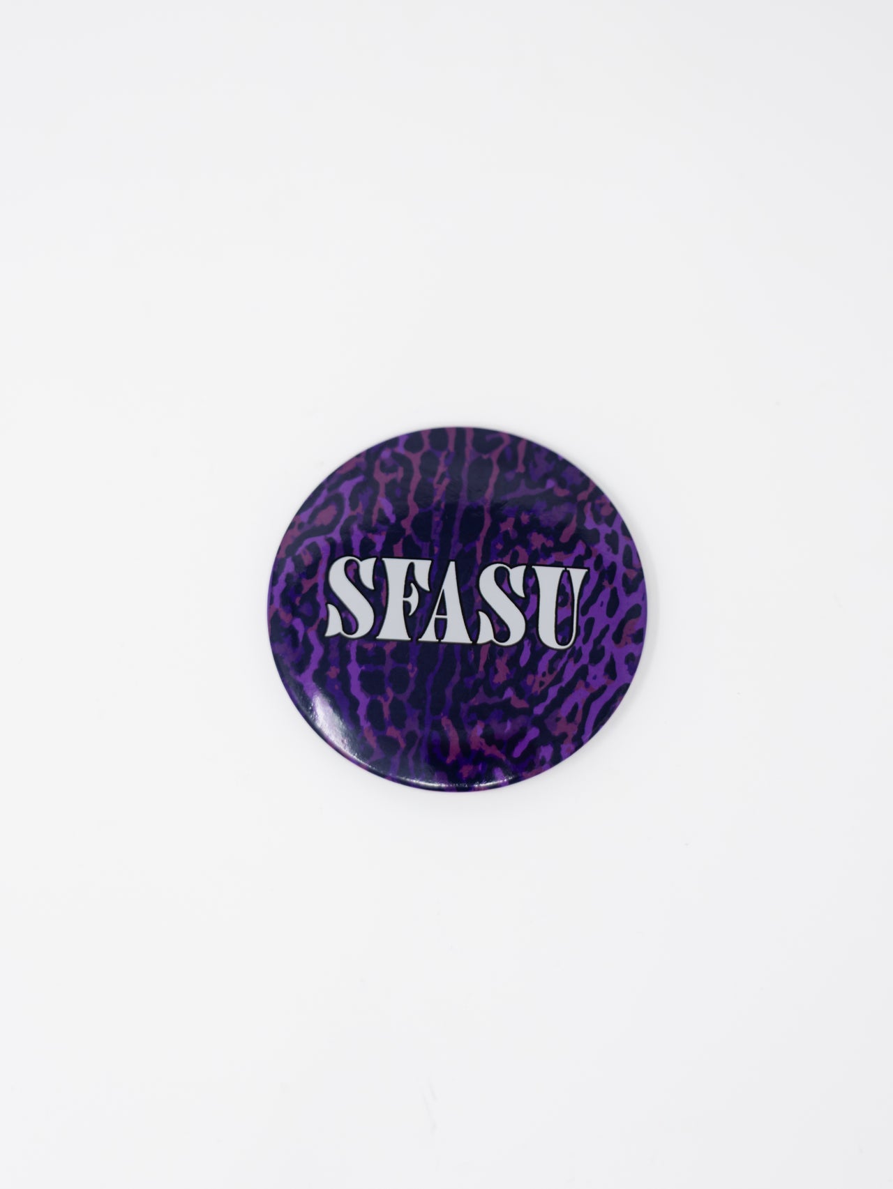 A round pin button featuring a dark purple and black leopard print background with "SFASU" printed across the center in bold white serif letters, displayed against a solid white background.