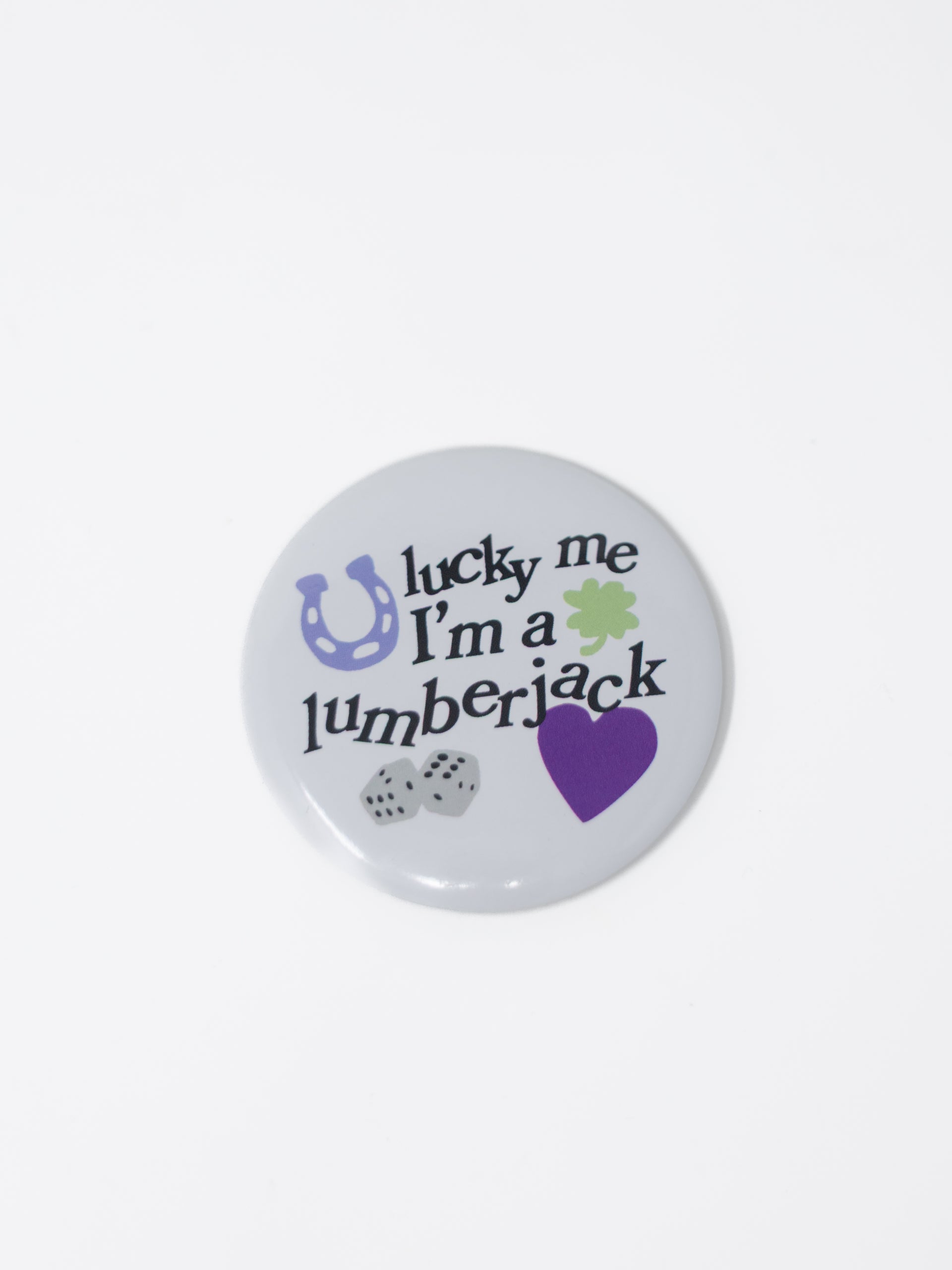 A round pin-back button badge with a light gray background and the text 'lucky me I'm a lumberjack' in black script. Decorative icons include a purple horseshoe, a green shamrock, two gray dice, and a purple heart.