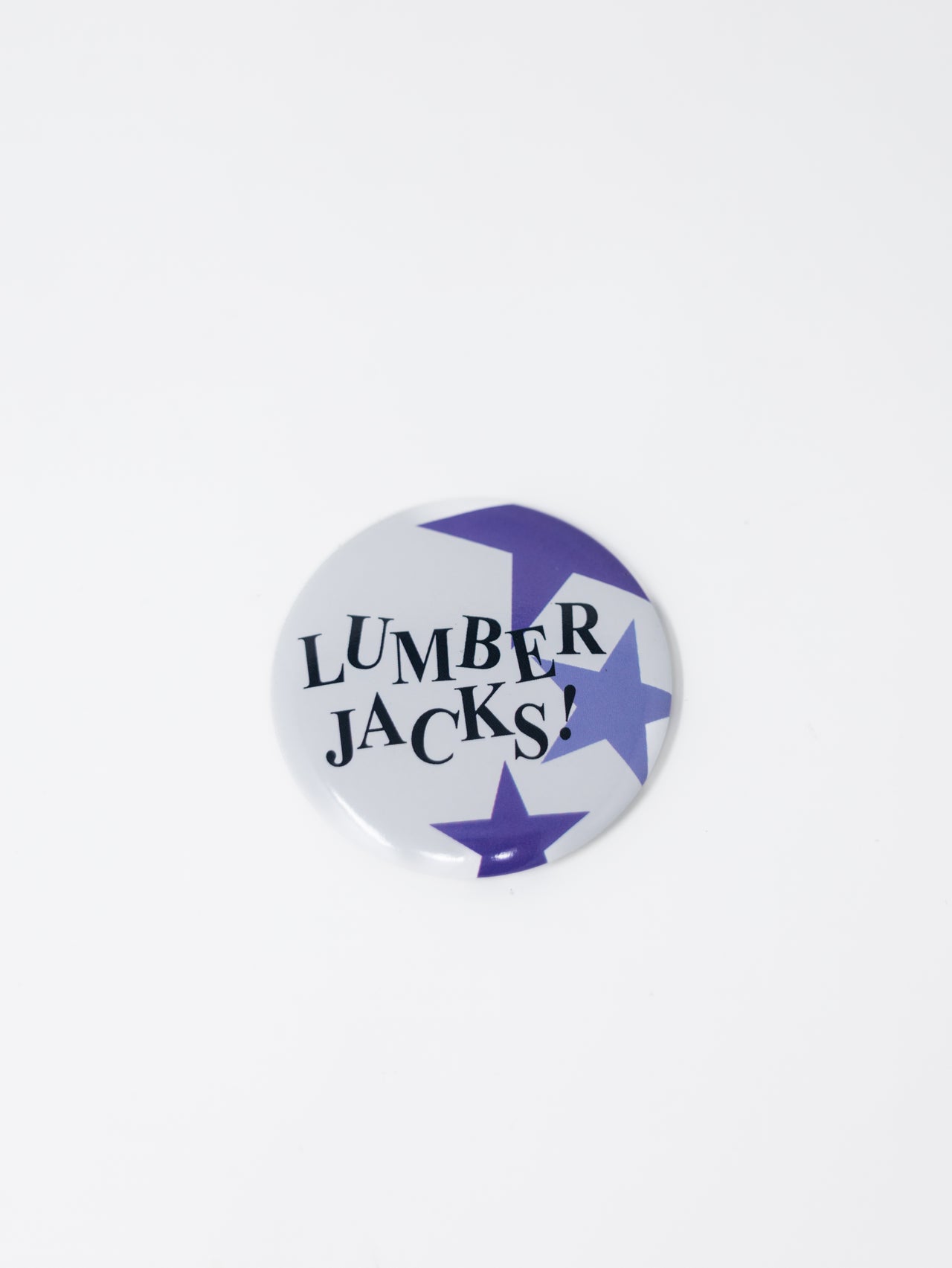 A round pin button featuring a white background with "LUMBERJACKS!" written in bold black serif text on the left, decorated with vibrant purple stars on the right and bottom, set against a plain white background.