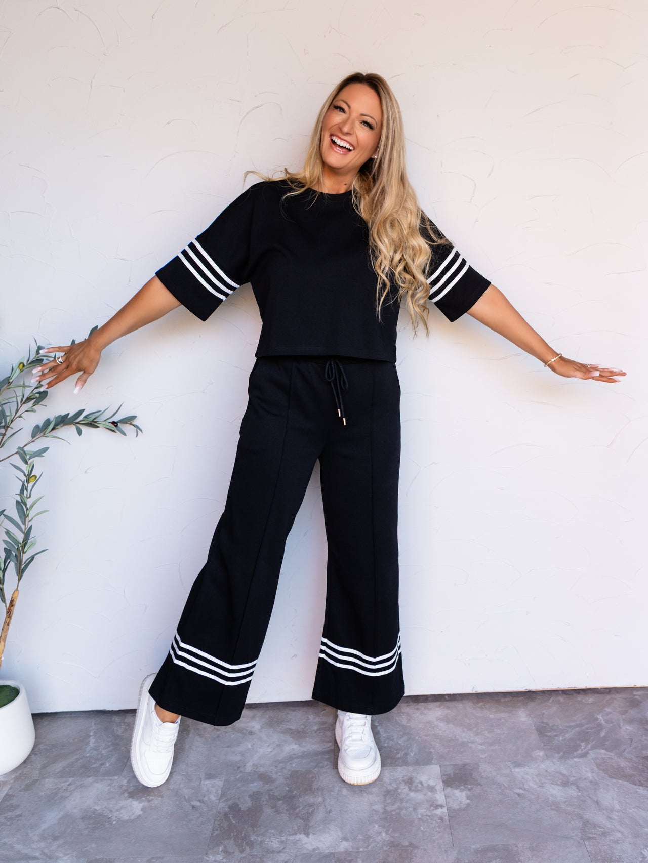 A full-length photograph of a laughing woman with long blonde wavy hair posing playfully with her arms open wide and leaning to one side against a textured white wall. She is wearing a black matching two-piece loungewear set featuring white athletic stripes on the short sleeves and wide-leg pant cuffs, styled with white platform sneakers.