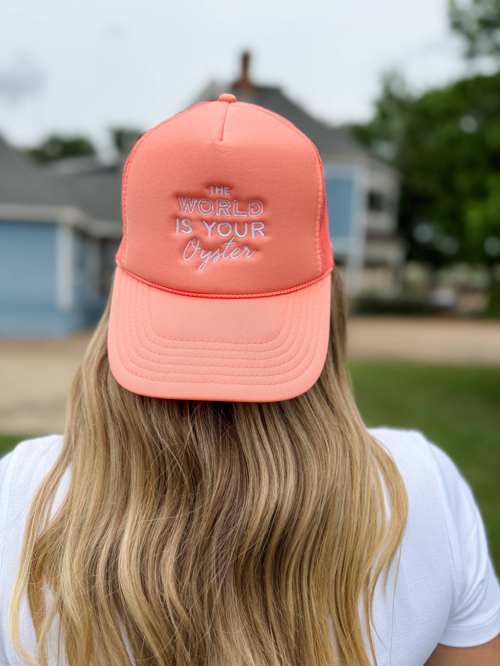 Person wearing a coral cap with 'The World is Your Oyster' text, outdoors.