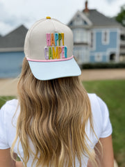 Person wearing a cap with 'Happy Camper' text in colorful letters, standing outdoors.
