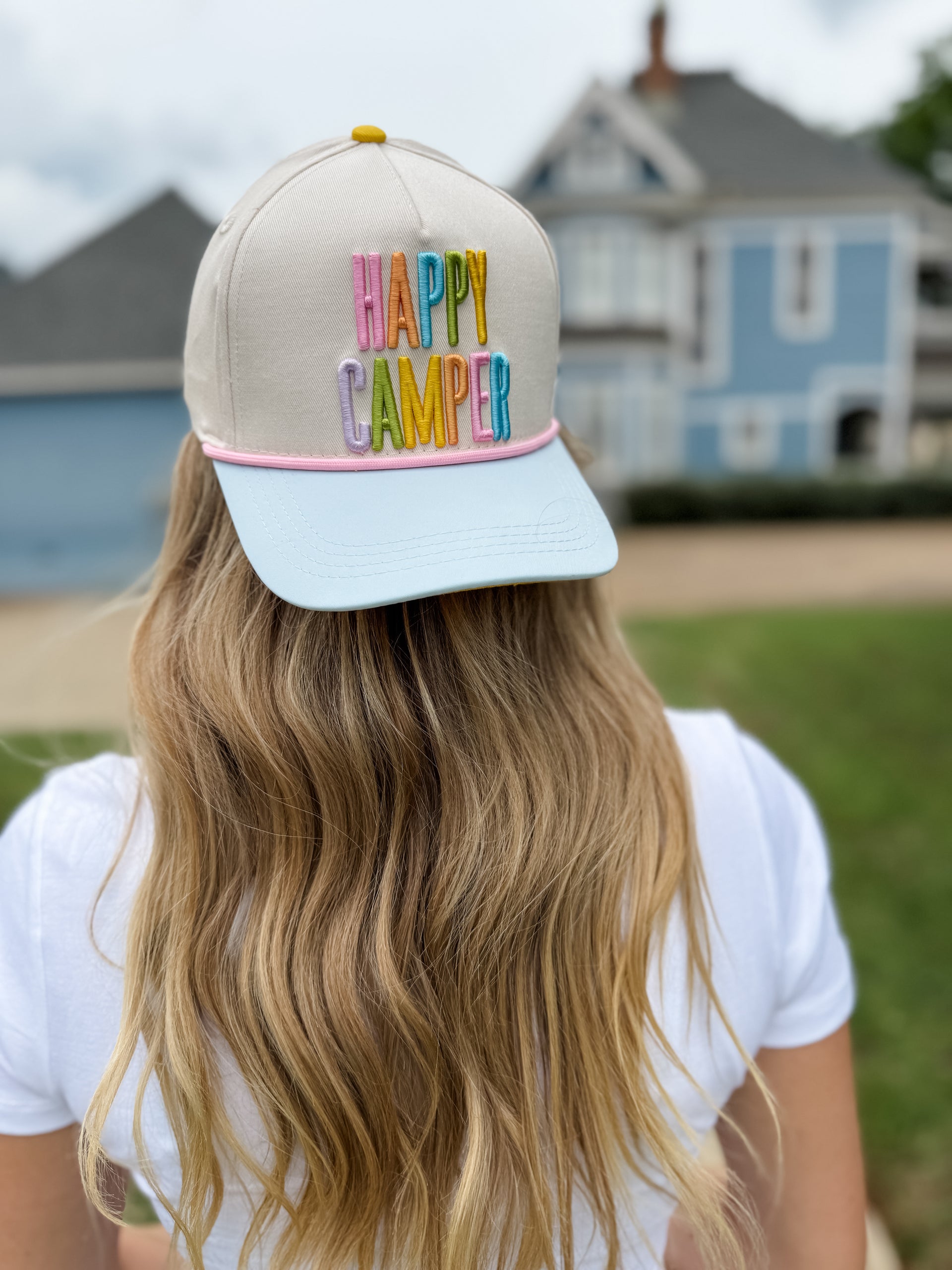 Person wearing a cap with 'Happy Camper' text in colorful letters, standing outdoors.