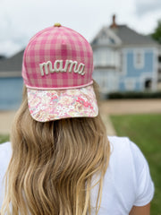 Person wearing a pink checkered cap with 'mama' on top, floral cap underneath, in front of a house.
