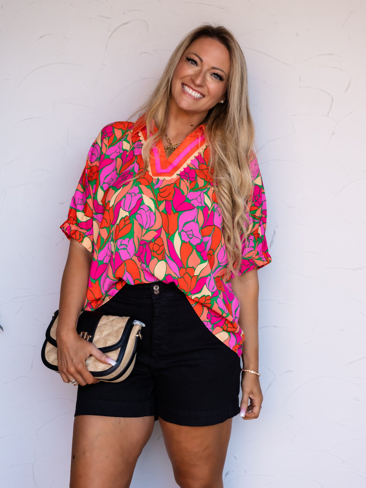A portrait photograph focusing from the mid-thigh up of a smiling woman with long blonde wavy hair posing against a textured white wall. She is wearing a short-sleeved blouse featuring a bold abstract floral print in bright pink, orange, and green made from 100% polyester fabric, designed with a collared V-neckline adorned with orange and pink ric-rac trim, styled with high-waisted black denim shorts, a gold statement necklace, and holding a straw crossbody clutch bag.