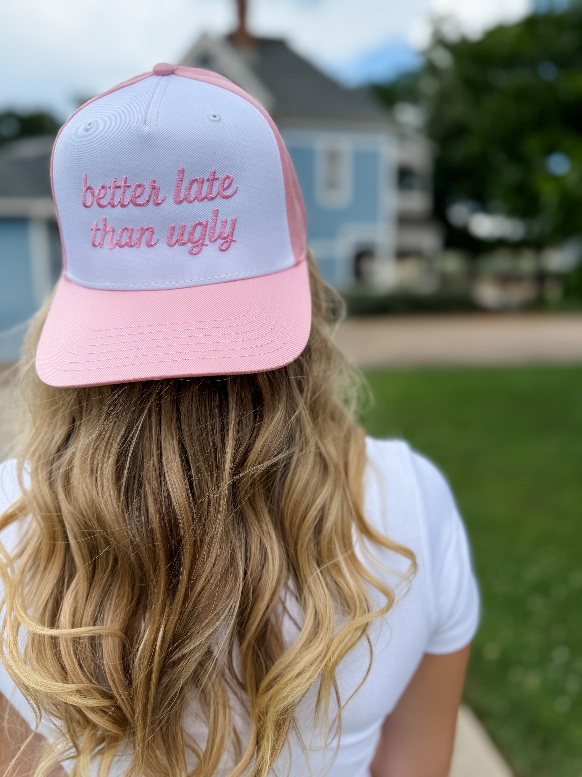 Person wearing a pink and gray cap with 'better late than ugly' text outdoors.