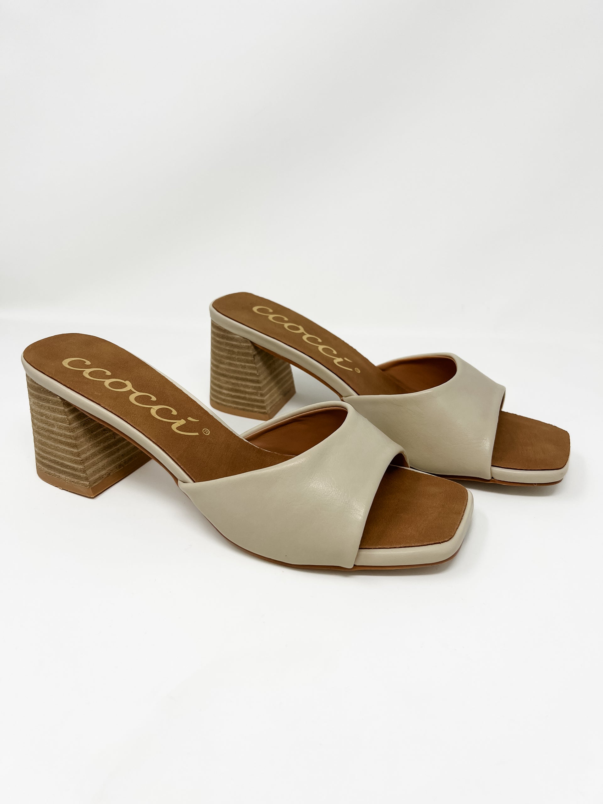 A pair of Ccocci brand heeled slide sandals displayed against a solid white background. The sandals feature a single, wide cream-colored faux leather strap over the foot, a square-toe silhouette, and a brown faux leather footbed with the brand name "Ccocci" printed in gold cursive. They are supported by a chunky, textured wood-grain style block heel.