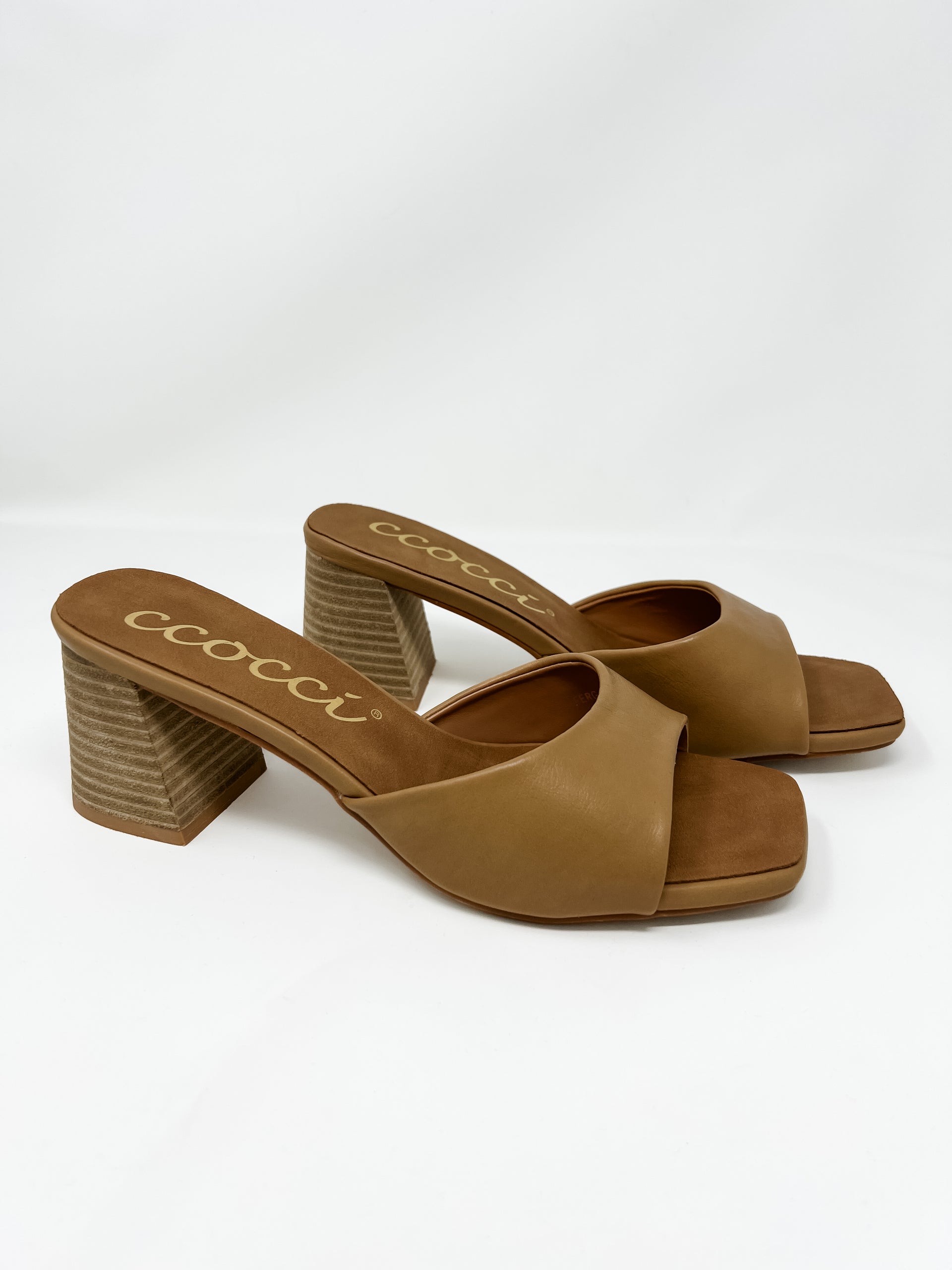 A pair of Ccocci brand heeled slide sandals in a warm camel tan color, displayed against a clean white background. The sandals feature a single, wide camel faux leather strap over the foot, a matching brown faux suede footbed with the gold cursive "Ccocci" brand logo, a modern square-toe front, and a chunky, wood-grain textured block heel.