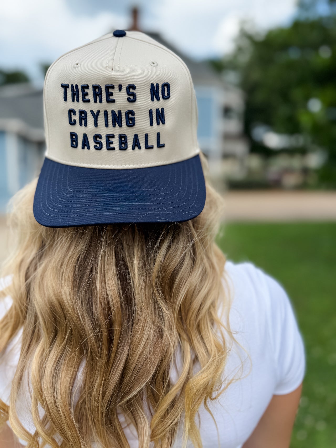 Person wearing a cap with 'There's no crying in baseball' text, blurred outdoor background