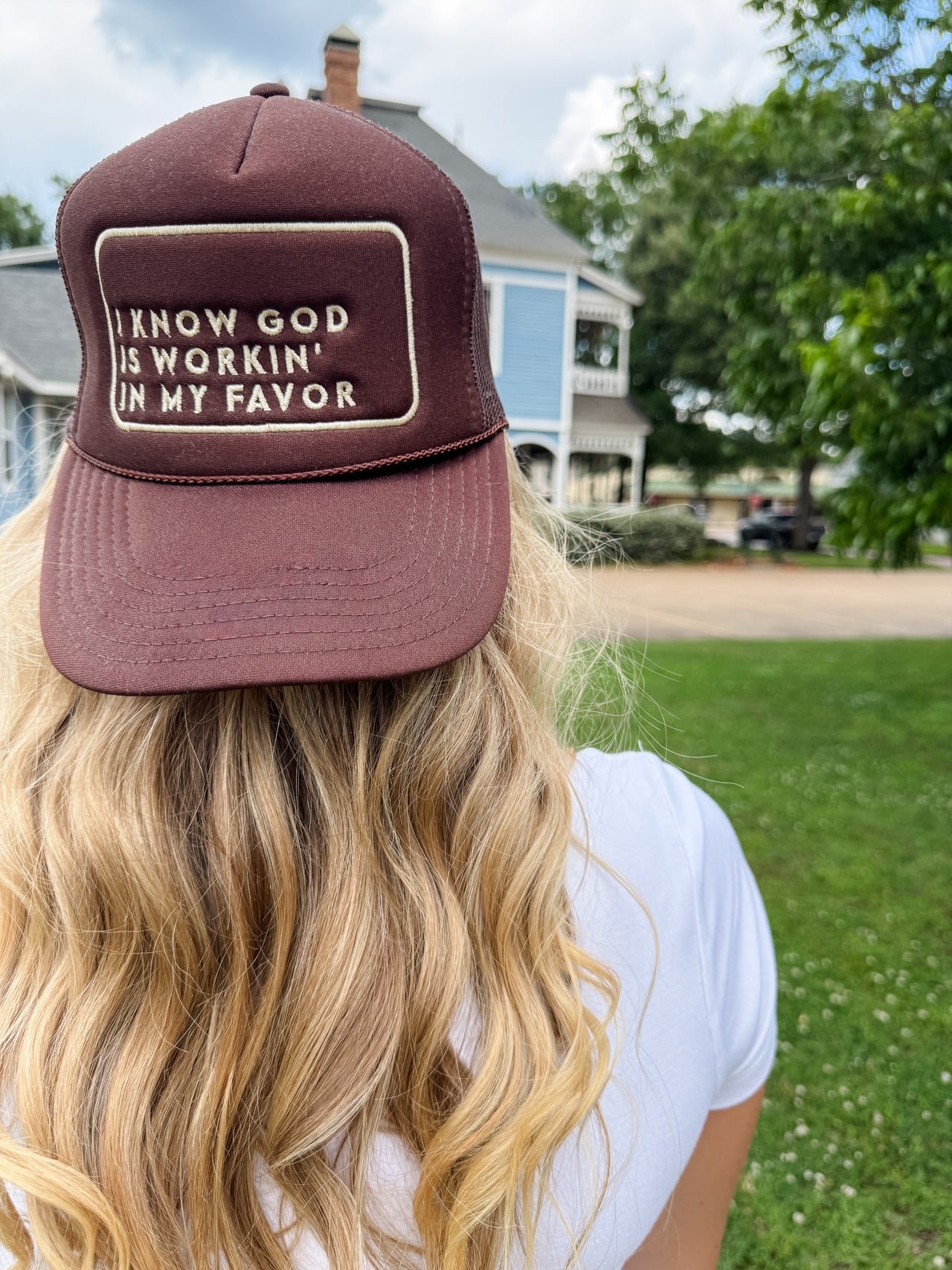 Person wearing a maroon cap with text, standing outdoors with a house and trees in the background