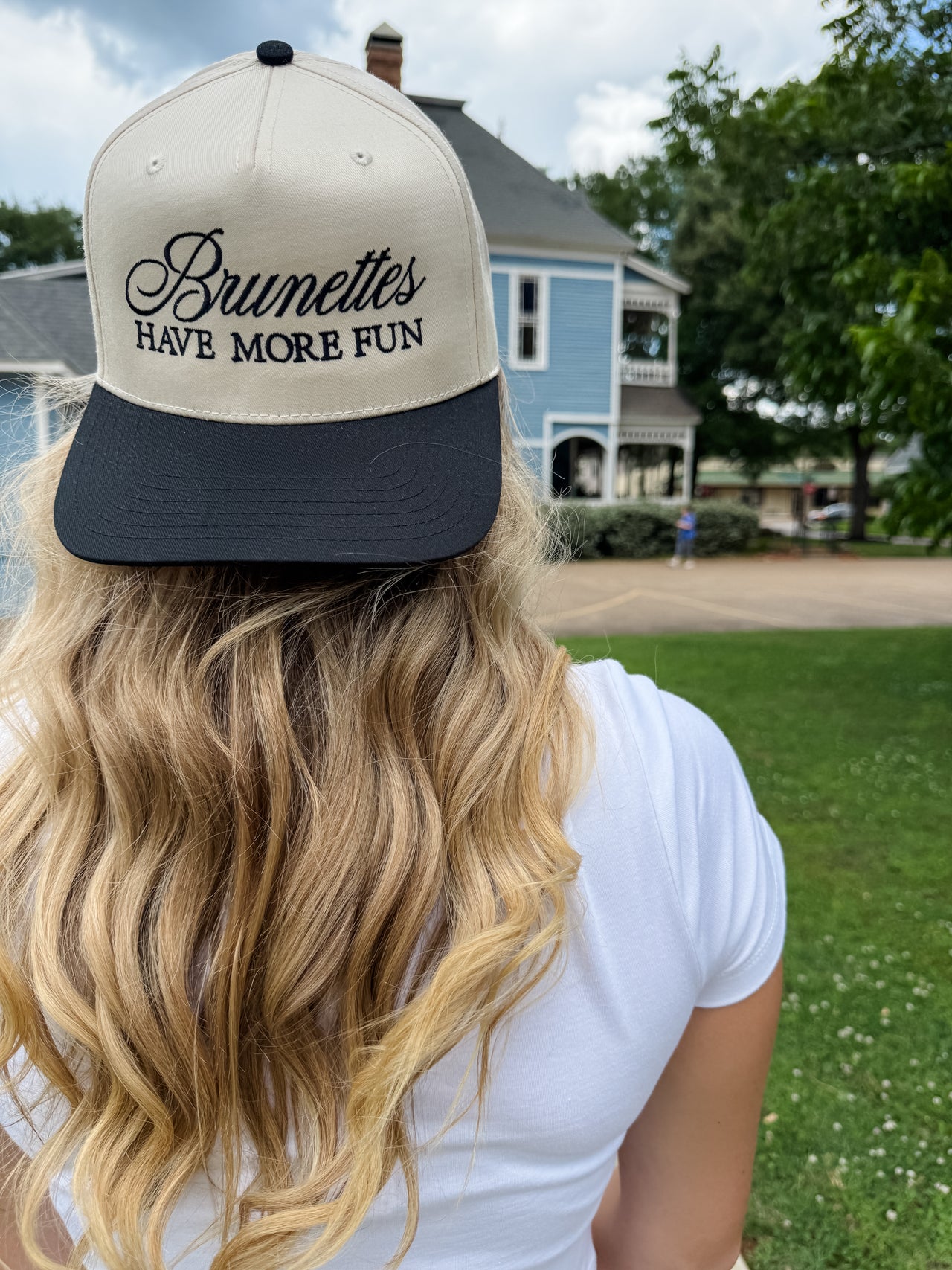 Person wearing a cap with 'Brunettes Have More Fun' text, standing outdoors with a house and trees in the background.