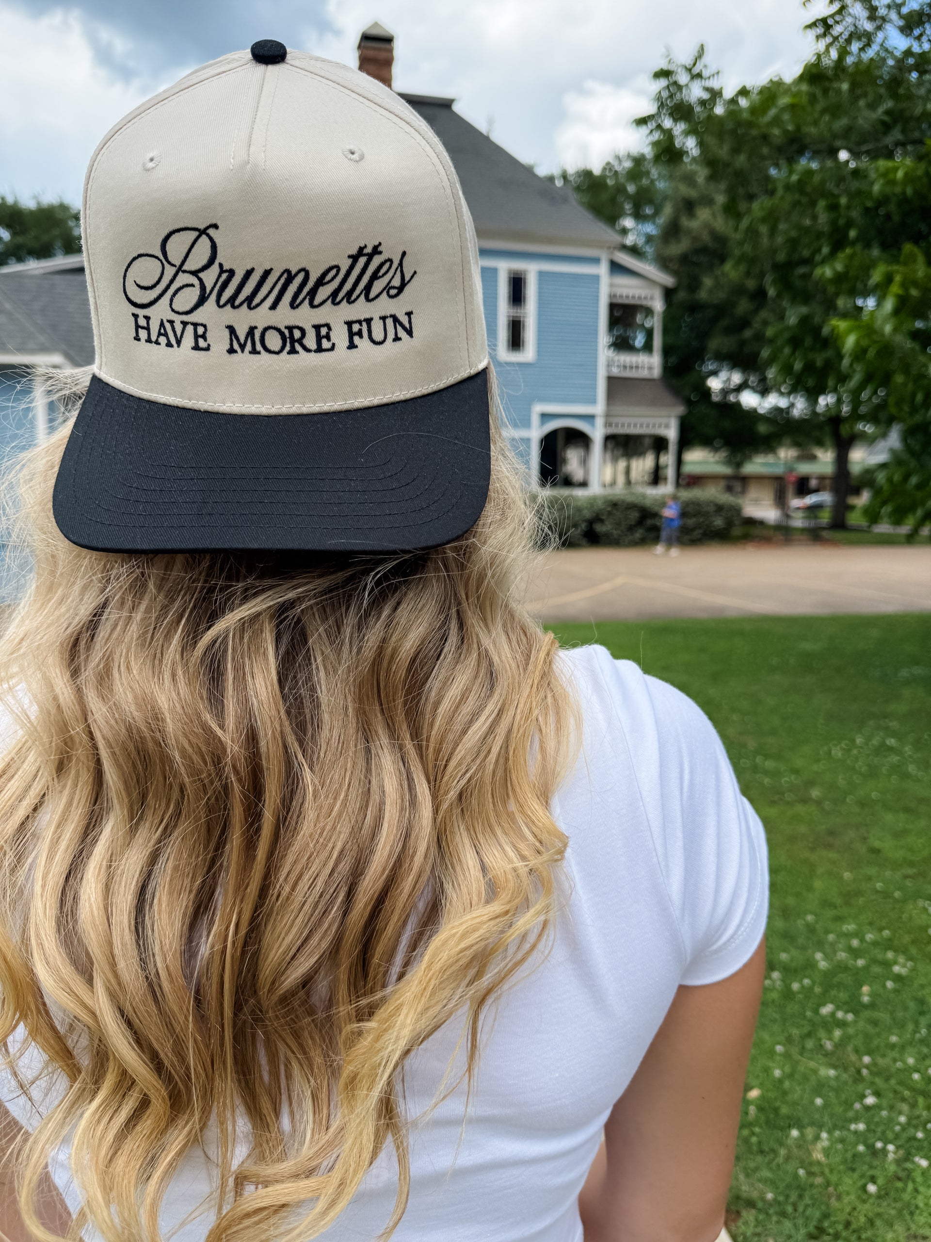 Person wearing a cap with 'Brunettes Have More Fun' text, standing outdoors with a house and trees in the background.