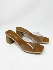 A pair of Ccocci brand heeled slide sandals with a transparent strap, shown against a solid white background. The sandals feature a clear vinyl top strap, a brown faux suede footbed with cream trim and the gold cursive "Ccocci" logo, a modern square toe, and a chunky, textured wood-grain block heel.