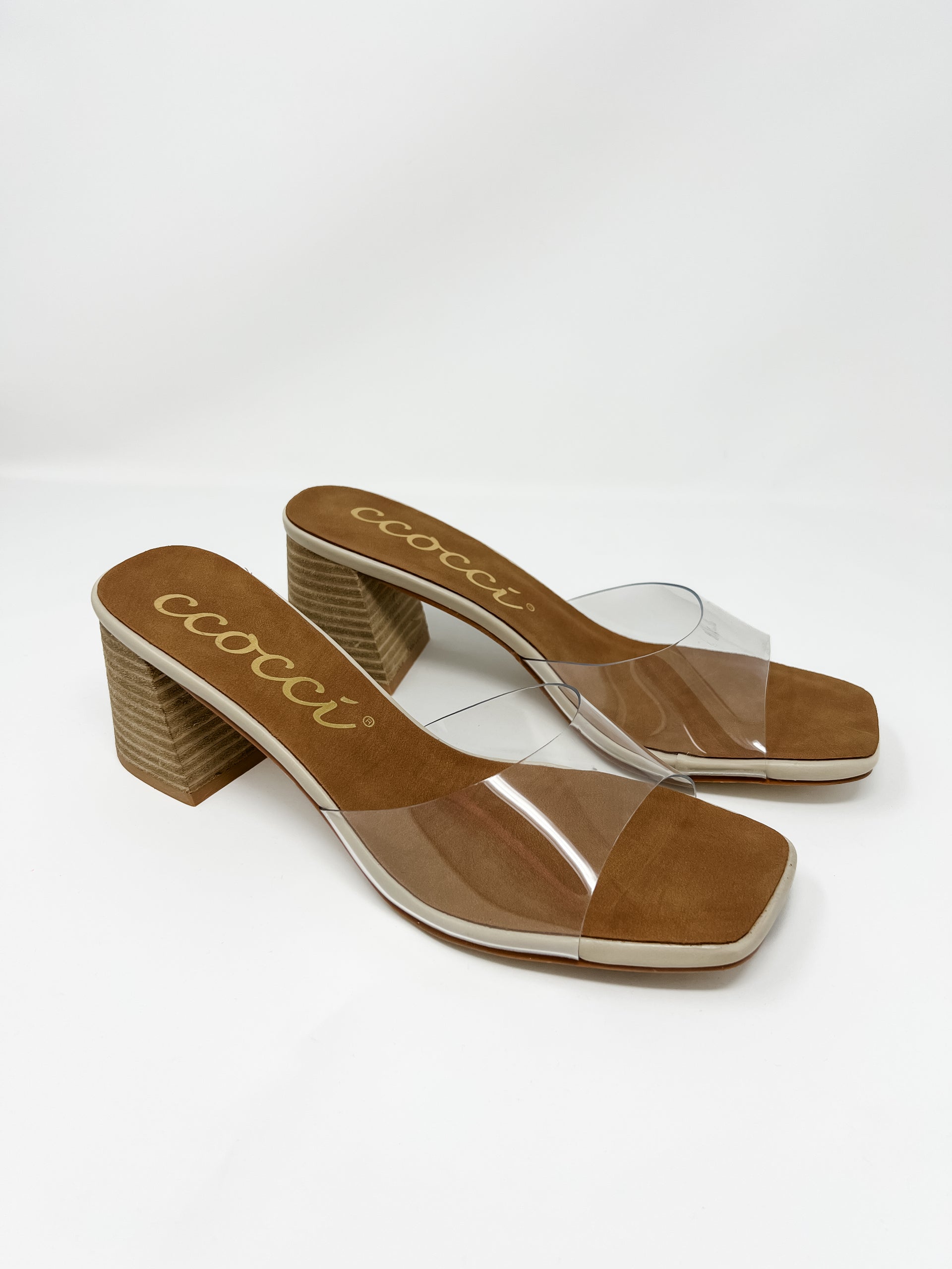 A pair of Ccocci brand heeled slide sandals with a transparent strap, shown against a solid white background. The sandals feature a clear vinyl top strap, a brown faux suede footbed with cream trim and the gold cursive "Ccocci" logo, a modern square toe, and a chunky, textured wood-grain block heel.