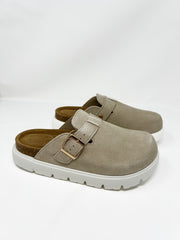 A pair of Project Cloud brand taupe-colored faux-suede platform clogs displayed against a plain white background. The slip-on shoes feature a closed, rounded toe, an adjustable instep strap with a shiny gold metal buckle, a contoured dark tan footbed with a cork-wrapped midsole, and a thick, chunky white platform lug sole.