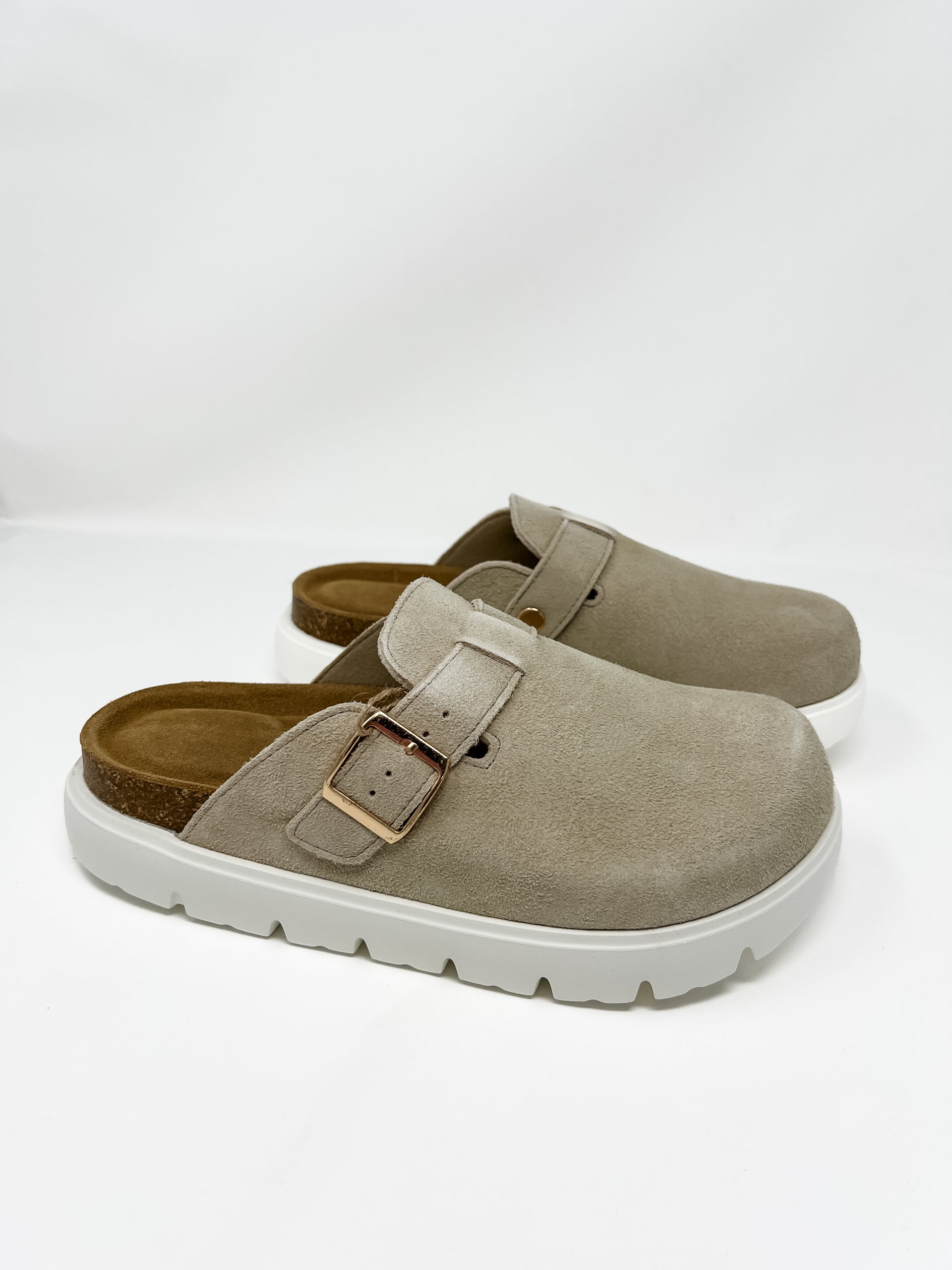 A pair of Project Cloud brand taupe-colored faux-suede platform clogs displayed against a plain white background. The slip-on shoes feature a closed, rounded toe, an adjustable instep strap with a shiny gold metal buckle, a contoured dark tan footbed with a cork-wrapped midsole, and a thick, chunky white platform lug sole.
