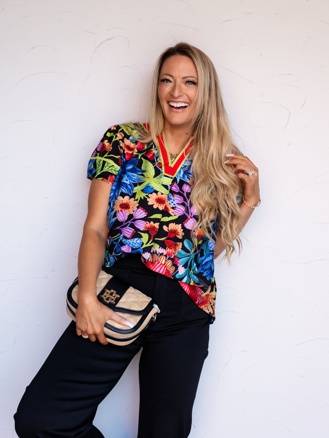 A portrait photograph focusing from the mid-thigh up of a laughing woman with long blonde wavy hair touching her hair and posing against a textured white wall. She is wearing a short-sleeved black blouse featuring a vibrant multicolored tropical botanical print made from an 80% rayon and 20% nylon blend fabric, designed with a V-neckline bordered in bright red and lime green trim, styled with black trousers, a gold statement necklace, and holding a straw crossbody clutch bag.
