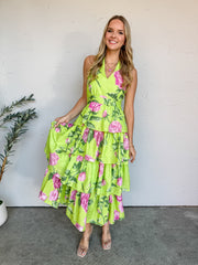 A close-up shot of a person standing against a textured off-white wall, wearing a lime green maxi dress crafted from a 100% cotton eyelet fabric featuring an all-over pink rose floral print. The dress showcases a tailored halter V-neckline, a defined waistband, and a voluminous, multi-tiered skirt detailing small embroidered eyelet cutouts throughout. She is styled with a simple gold link chain necklace, a thin gold bracelet, and clear, nude strappy block heels while standing on a concrete floor next to a p