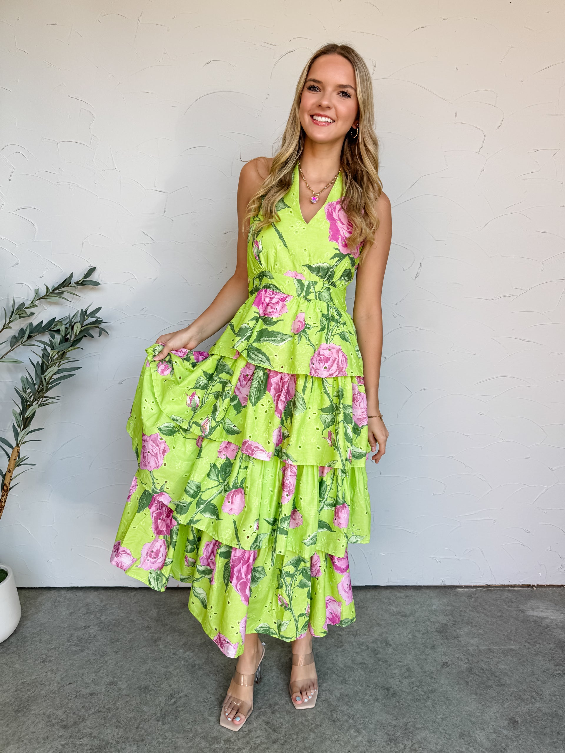 A close-up shot of a person standing against a textured off-white wall, wearing a lime green maxi dress crafted from a 100% cotton eyelet fabric featuring an all-over pink rose floral print. The dress showcases a tailored halter V-neckline, a defined waistband, and a voluminous, multi-tiered skirt detailing small embroidered eyelet cutouts throughout. She is styled with a simple gold link chain necklace, a thin gold bracelet, and clear, nude strappy block heels while standing on a concrete floor next to a p