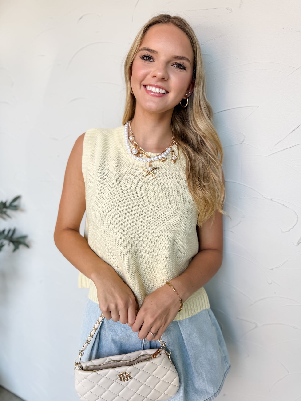 A woman wearing a buttercream yellow sleeveless vest made of a 100% acrylic knit construction, featuring a round crew neckline, a finely textured body, and a ribbed hemline. The top showcases a dramatic open-back design worn over a tan tube top, complete with a self-tie closure at the nape of the neck with long dangling knit straps framed by vertical ribbing. She is styled against a textured off-white wall wearing light-wash denim shorts, a chunky gold chain and pearl necklace with a large starfish charm, a