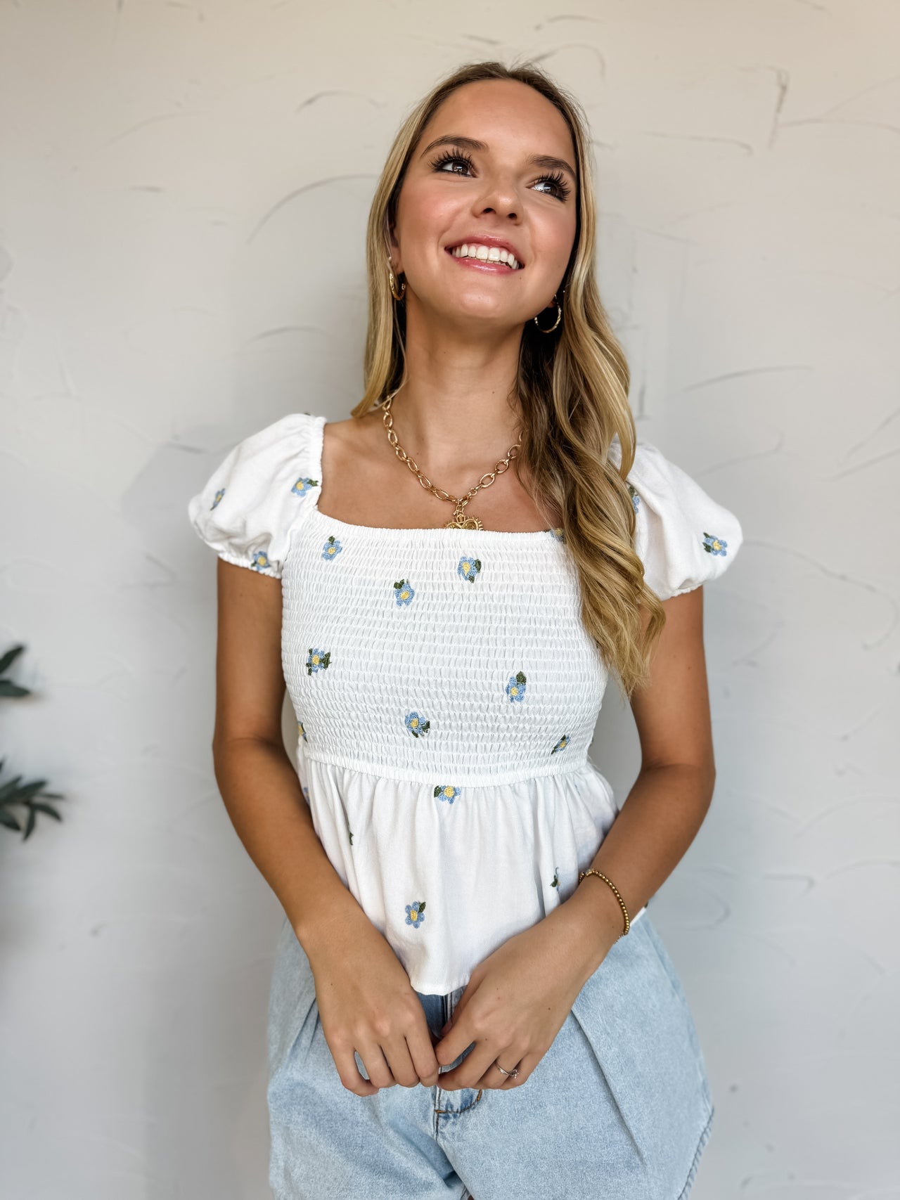 Smiling model wearing a white embroidered peplum top featuring a square neckline, fully smocked bodice, short puff sleeves with elasticized cuffs, and delicate blue floral embroidery throughout. The lightweight woven top has a feminine peplum silhouette that flows from the waist. Styled with light wash denim shorts, a gold heart pendant necklace, and a woven straw shoulder bag with a gold chain strap against a neutral studio backdrop.