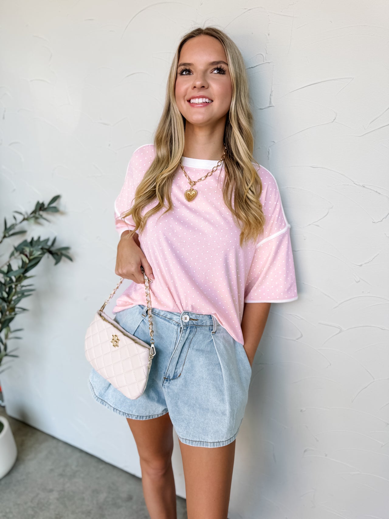 Smiling model wearing a relaxed-fit blush pink top made from a soft cotton-spandex knit fabric. The top features an allover white polka dot print, crew neckline, dropped shoulders, short sleeves, and white contrast binding at the neckline, sleeve cuffs, and hemline. The lightweight fabric has a comfortable feel with slight stretch. Styled with distressed light wash denim shorts, layered gold necklaces, gold hoop earrings, and loose wavy hair. Photo is taken against a neutral studio background with the model