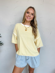 Smiling model wearing a relaxed-fit Yellow top made from a soft cotton-spandex knit fabric. The top features an allover white polka dot print, crew neckline, dropped shoulders, short sleeves, and white contrast binding at the neckline, sleeve cuffs, and hemline. The lightweight fabric has a comfortable feel with slight stretch. Styled with distressed light wash denim shorts, layered gold necklaces, gold hoop earrings, and loose wavy hair. Photo is taken against a neutral studio background with the model